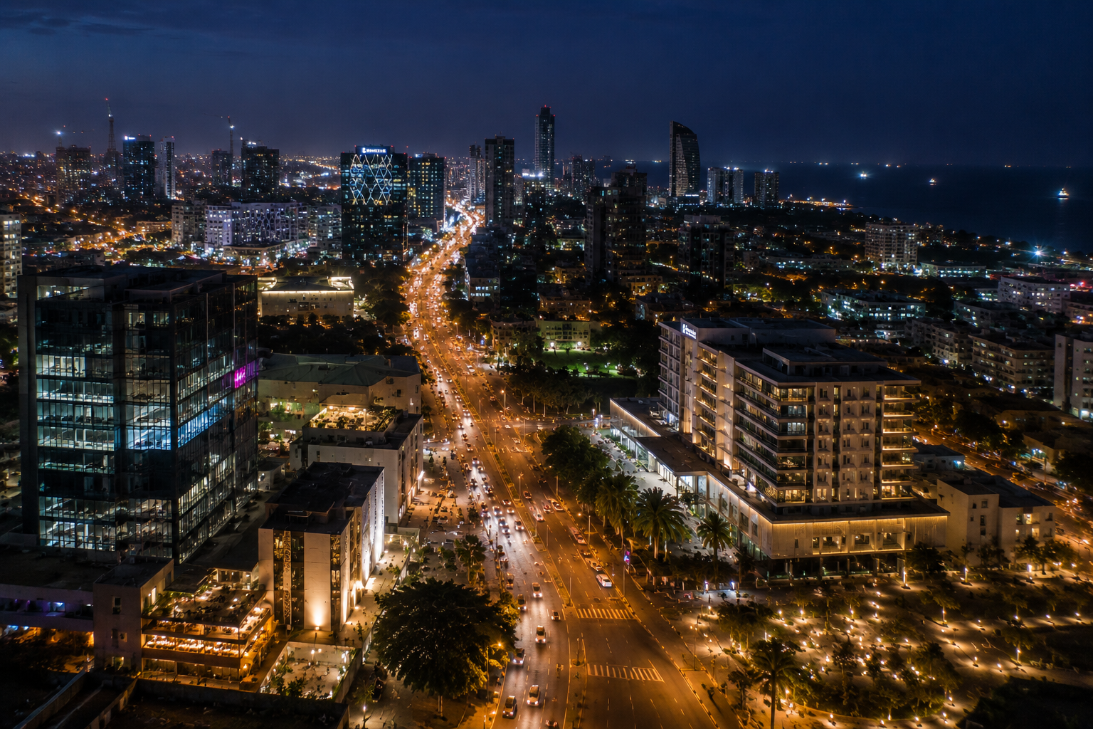 A city boulevard at night, traffic light-trails running between lit towers toward the sea.